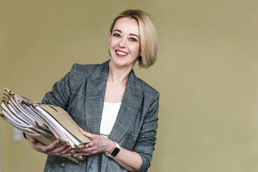 Smiling blond businesswoman with pile of documents in front of wall - OGF01052