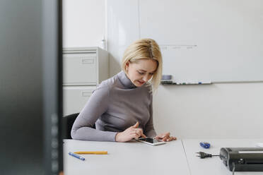 Blonde Geschäftsfrau, die ein Smartphone benutzt, sitzt am Schreibtisch im Büro - OGF01067