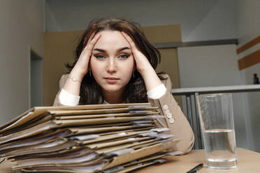 Tired young businesswoman sitting with hand in hair at desk in office - OGF01079