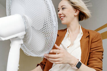 Lächelnde blonde Geschäftsfrau mit zerzaustem Haar sitzt vor einem elektrischen Ventilator - OGF01085