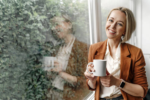 Glückliche blonde Frau, die eine Kaffeetasse hält und vor einem Glasfenster im Büro steht - OGF01086