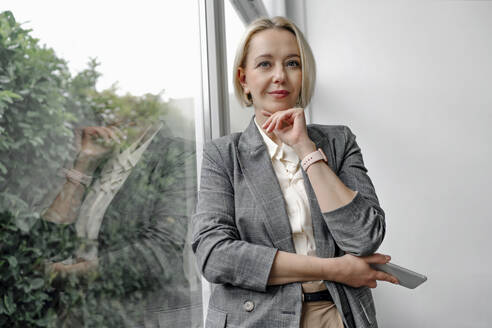 Beautiful blond businesswoman standing with hand on chin by glass window in office - OGF01087