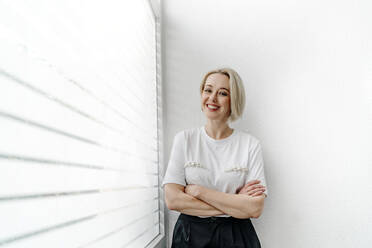 Cheerful blond businesswoman standing with arms crossed by window in office - OGF01089