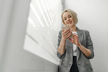 Blonde weibliche Fachkraft, die eine SMS über ihr Smartphone am Fenster im Büro schreibt - OGF01091