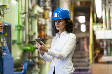 Weiblicher Ingenieur mit Schutzhelm, der ein Mobiltelefon benutzt, während er in der Industrie steht - MOEF03787