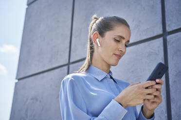 Geschäftsfrau mit drahtlosen In-Ear-Kopfhörern, die ihr Mobiltelefon an der Wand benutzt - ABIF01411