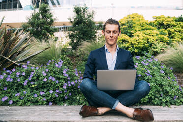 Smiling businessman using laptop while sitting with cross-legged on bench - ASGF00810