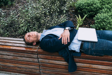 Businessman lying with laptop on bench at park - ASGF00818