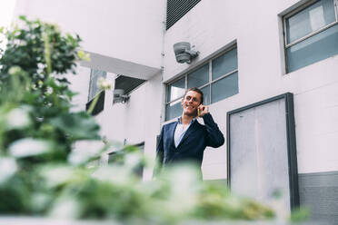Geschäftsmann lächelt, während er mit einem Smartphone vor einem Gebäude spricht - ASGF00825