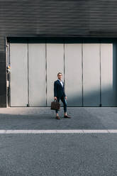 Businessman holding briefcase while standing on footpath - ASGF00828