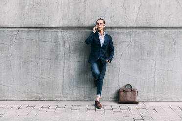Businessman talking on mobile phone while standing by briefcase on footpath - ASGF00837