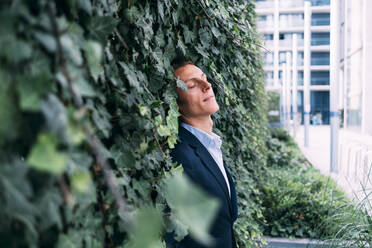 Businessman with eyes closed leaning on plant wall - ASGF00843