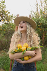 Positive Frau mit geöffnetem Mund, stehend mit Zitrusfrüchten in den Händen in einem Obstgarten während der Erntesaison - ADSF26967