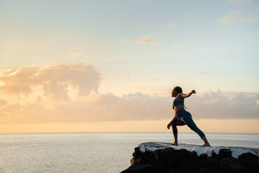 Seitenansicht der weiblichen Praxis Yoga auf Utthita Parsvakonasana Haltung auf Felsen gegen Meer unter bewölktem Himmel bei Sonnenuntergang - ADSF27037