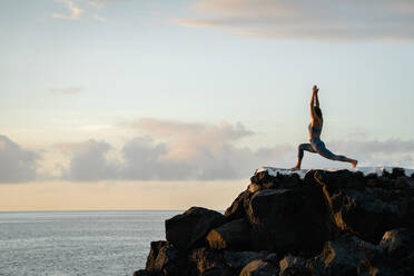 Seitenansicht einer anonymen Frau, die Yoga mit erhobenen Armen auf Felsbrocken gegen den Ozean unter bewölktem Himmel bei Sonnenuntergang praktiziert - ADSF27038