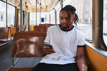 Junger positiver afroamerikanischer Mann in Freizeitkleidung, der im Zug der Wiener Bahn tagsüber in der Stadt auf seinem Handy surft - ADSF27042