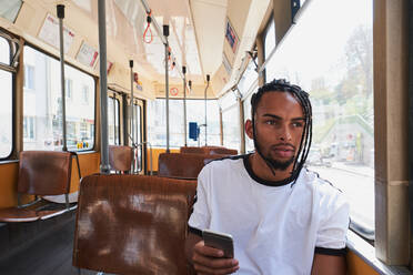 Junger positiver afroamerikanischer Mann in Freizeitkleidung, der im Zug der Wiener Bahn tagsüber in der Stadt auf seinem Handy surft - ADSF27043