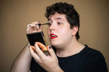 Konzentrierter, exzentrischer, weiblicher Mann, der Wimperntusche mit einer Bürste aufträgt, während er sich mit geöffnetem Mund schminkt und einen Spiegel hält - ADSF27055