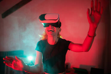 Happy woman wearing t shirt while using VR goggles and standing in studio with red neon lights - ADSF27058