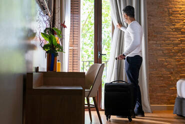 Full body low angle side view of adult man with luggage opening curtain hanging on balcony door in hotel room - ADSF27061