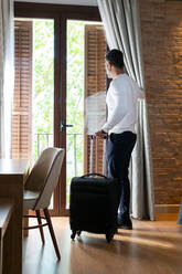 Full body low angle side view of adult man with luggage opening curtain hanging on balcony door in hotel room - ADSF27062