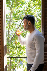 Crop männlich bärtigen Mann trinken Orangensaft aus dem Glas auf dem Balkon in sonnigen Tag - ADSF27063