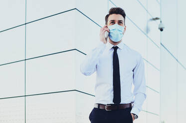 Stylish male executive in formal clothes and medical mask talking on cellphone while looking away in town - ADSF27071