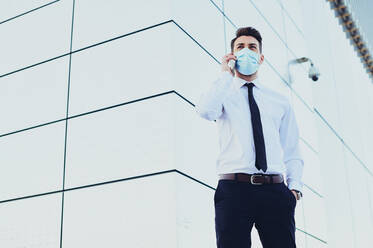 Stylish male executive in formal clothes and medical mask talking on cellphone while looking away in town - ADSF27072