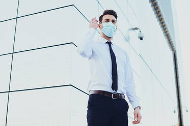 From below of male entrepreneur in formal wear with medical mask looking away in town during coronavirus pandemic - ADSF27073