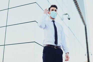 Von unten: männlicher Unternehmer in formeller Kleidung mit medizinischer Maske, der in der Stadt während der Coronavirus-Pandemie wegschaut - ADSF27073