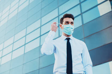 From below of male entrepreneur in formal wear with medical mask looking away in town during coronavirus pandemic - ADSF27074