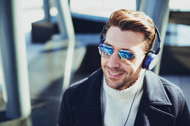 Lächelnder, bärtiger Mann mit moderner Sonnenbrille, der in der Stadt im Gegenlicht einen Song über Kopfhörer hört - ADSF27082