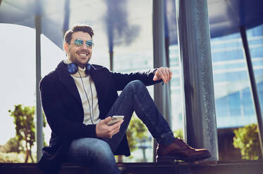 Content male in stylish apparel with headphones and cellphone sitting in city on sunny day - ADSF27084