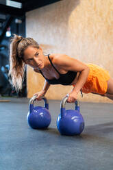 Entschlossene Sportlerin in Sportkleidung, die während eines Funktionstrainings mit Gewichten auf dem Boden im Fitnessstudio in der Plank-Position steht und wegschaut - ADSF27090