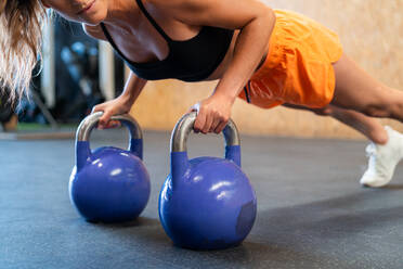 Abgeschnittene, nicht erkennbare, entschlossene Sportlerin in Sportkleidung, die während des Funktionstrainings mit Gewichten auf dem Boden im Fitnessstudio in Plank-Pose steht - ADSF27091