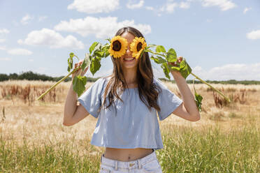 Frau bedeckt Augen mit Sonnenblumen, während sie auf einer Wiese steht - EIF01635