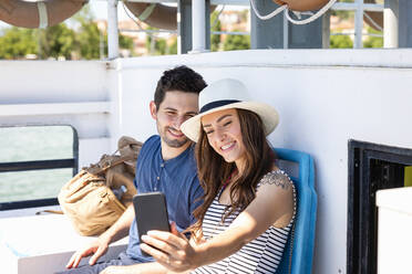 Smiling woman taking selfie with man through mobile phone while sitting in tourboat - EIF01654