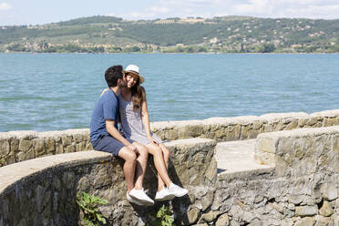 Man kissing smiling girlfriend while sitting on retaining wall - EIF01678