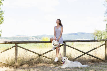 Smiling woman holding hat and pet leash while sitting on railing - EIF01682