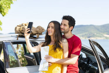 Lächelndes Paar, das ein Selfie mit seinem Smartphone macht, während es am Auto steht - EIF01699