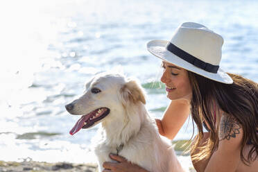 Smiling woman with hat stroking dog at lakeshore - EIF01715
