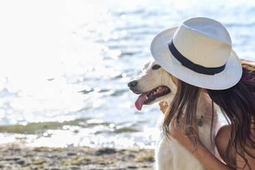 Woman wearing hat kissing dog at lakeshore on sunny day - EIF01716