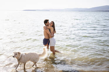 Smiling couple embracing while standing by dog in lake - EIF01720