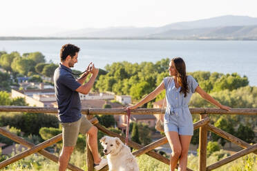 Man photographing woman through camera while standing with dog by railing - EIF01730