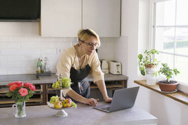 Female professional wearing apron using laptop in kitchen - KMKF01705