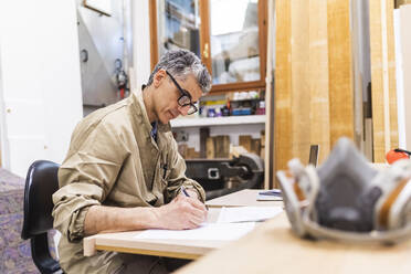 Älterer männlicher Schreiner, der am Tisch in der Werkstatt auf Papier schreibt - MEUF03540