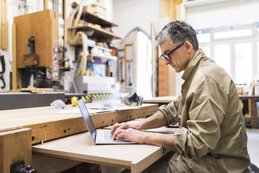 Älterer männlicher Tischler arbeitet in der Werkstatt am Laptop - MEUF03542