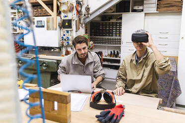Ein männlicher Schreiner benutzt einen Virtual-Reality-Simulator, während sein Kollege in der Werkstatt einen Laptop benutzt - MEUF03588