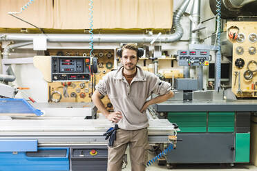 Junger männlicher Handwerker steht mit den Händen in den Hüften vor einer Maschine - MEUF03595