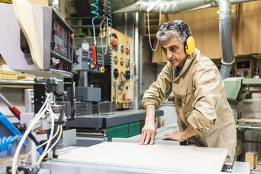 Mature male carpenter wearing ear protectors working on machinery at workshop - MEUF03596